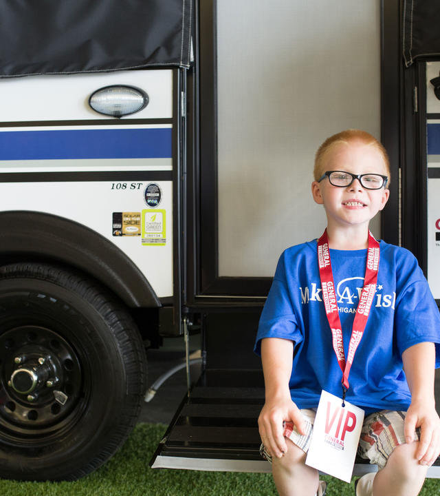 A child with short red hair and black glasses sits on the entry ramp to a camper. The child is wearing a royal blue Make-A-Wish t-shirt, tan plaid shorts, and a red lanyard with a large tag that says VIP.