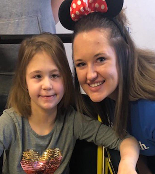 An adult with shoulder-length straight dark hair crouches next to a child sitting in a yellow wheelchair. The adult is wearing a royal blue Make-A-Wish t-shirt, jeans, and Minnie Mouse ears. The child is wearing a long-sleeved gray t-shirt with a multi-colored heart, jeans, and purple cowboy boots and clutching a large brown and white stuffed horse.
