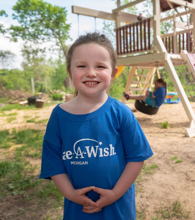 A young child in a royal blue Make-A-Wish t-shirt with brown hair pulled into a loose ponytail stands in front of a backyward playset with a wide smile. In the background, three siblings are playing on the swings.