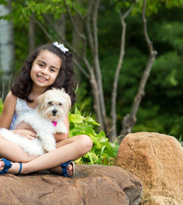 A 7-year-old child with shoulder-length curly dark hair cuddles a fluffy white puppy while sitting cross-legged on a large rock. The child is wearing a white lace tank top and shorts with blue sandals. In the background there are large green plants and trees.