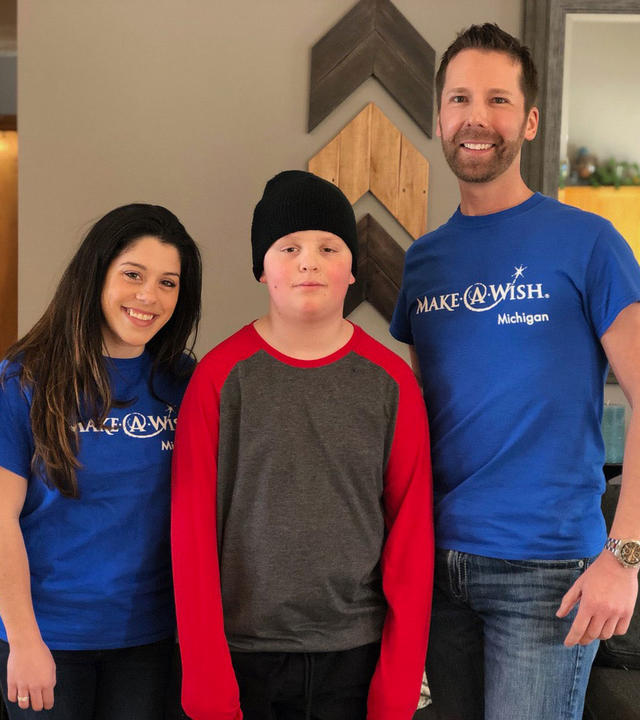 Two adults in royal blue Make-A-Wish t-shirts and jeans stand on either side of a child in a red and gray shirt and black beanie. The background is a living room with a gray couch and wooden wall art.