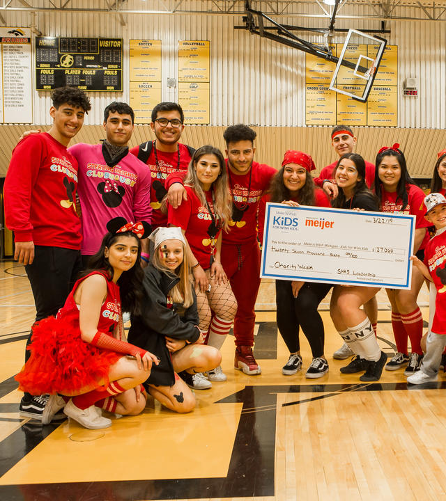 17 high school students wear red headbands and shirts that read "Sterling Heights Clubhouse" as they pose in their school gymnasium with a child. The group holds an oversized check for $27,060 dollars made out to Make-A-Wish Michigan.