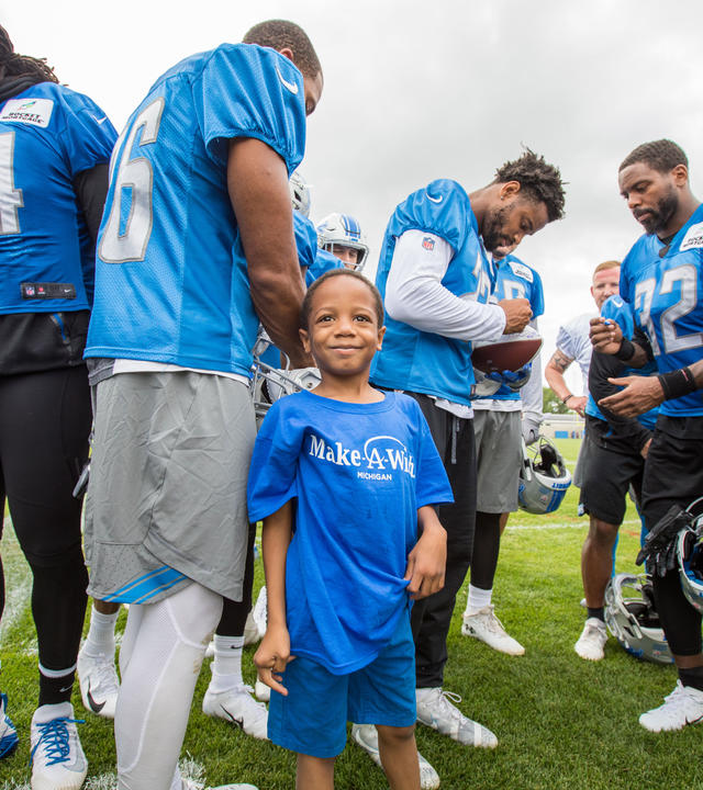 A child in a royal blue Make-A-Wish t-shirt and jean shorts stands amidst a crowd of football players wearing practice gear. The child is smiling at the camera, while the players are passing around and signing a football.