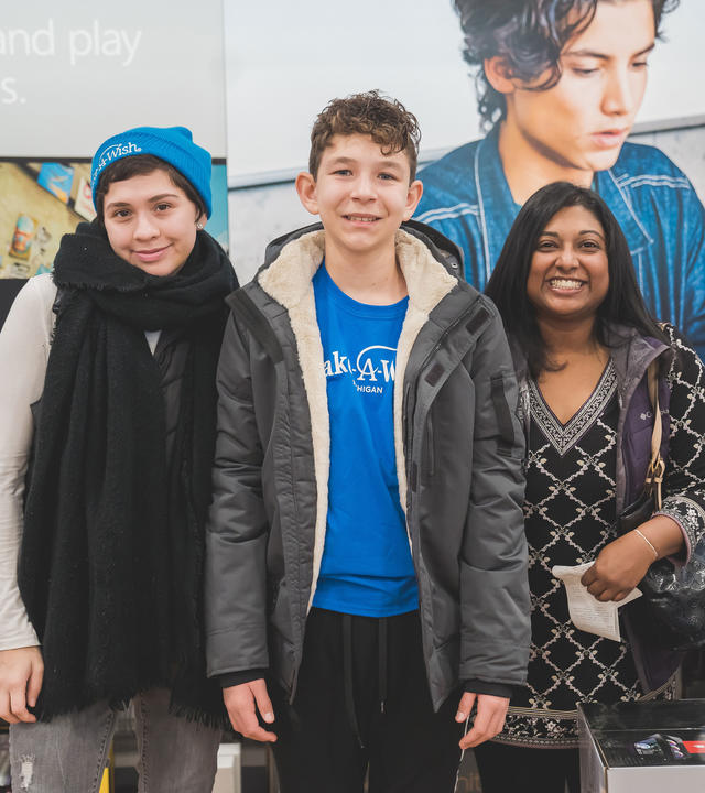 A teenagee with short curly brown hair, wearing a royal blue Make-A-Wish t-shirt, gray winter jacket and black athletic pants, smiles broadly. The teenager stands between two wish-granting volunteers in Best Buy's computer department. On the left, volunteer Timerra is wearing a royal blue Make-A-Wish beanie and a long black scarf. On the right, volunteer Millie is wearing a black and white patterned tunic and holding the receipt from the purchases made at Best Buy. 