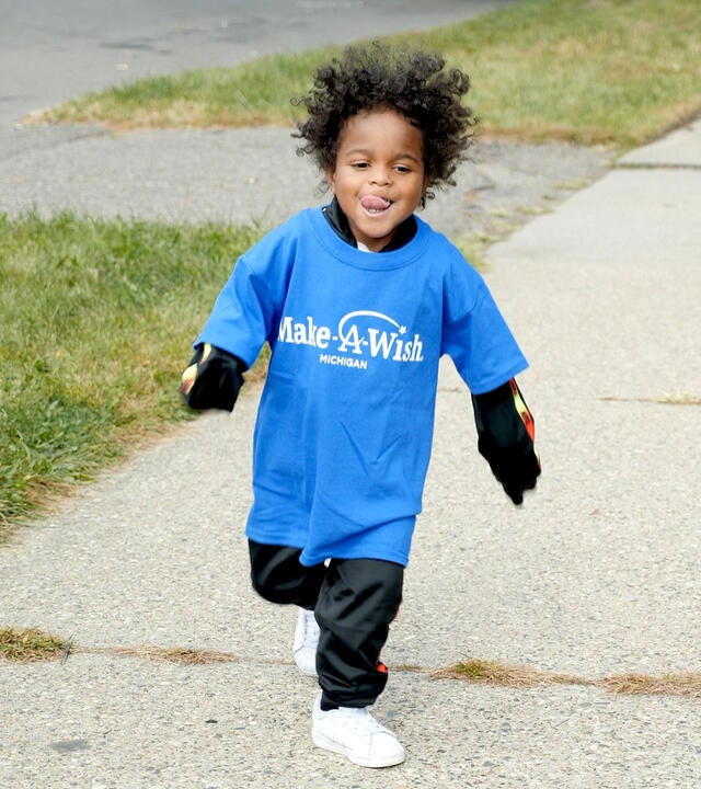 A young child with curly dark hair runs happily down a sidewalk. The child is wearing a royal blue Make-A-Wish t-shirt over a black long-sleeve shirt, black pants and white tennis shoes.