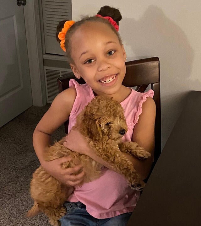 A child wearing a sleeveless pink shirt and jeans sits in a chair holding a small, fluffy golden puppy. The child's hair is done in two pigtail buns secured with bright scrunchies. There is a big smile on the child's face.