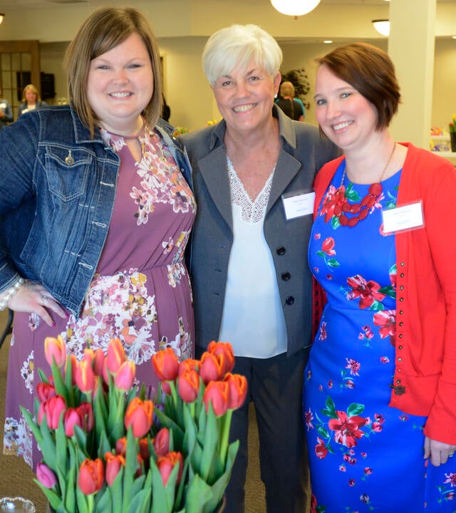 Three adults in bright clothing stand with their arms around each other. In front of them is a large bouquet of red tulips.