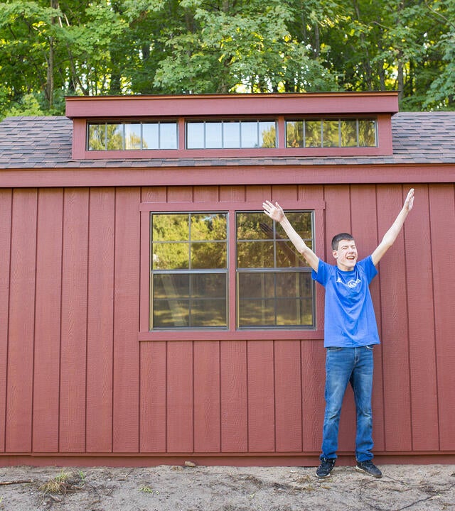 A teenage wish kid wearing a royal blue Make-A-Wish t-shirt and jeans stands in front of a red shed with a big smile and arms thrown high into the air in excitement.
