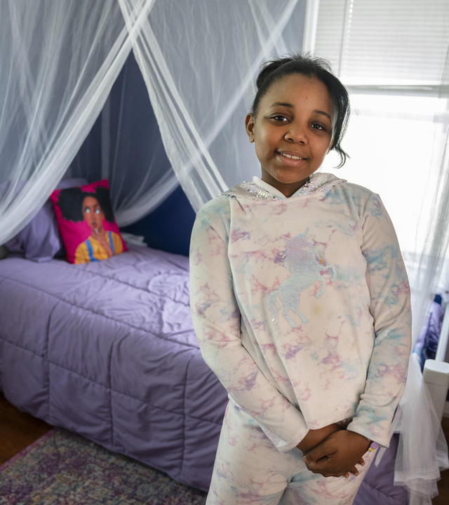 A child wearing a pastel tie-dyed hoodie with a unicorn design and matching pants stands with hands clasped in front of a bed decorated with sheet white curtains and a purple bedspread.