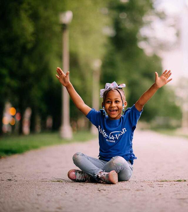 Taylor in Make-A-Wish blue tshirt, sitting on sidewalk with arms raised in joy.