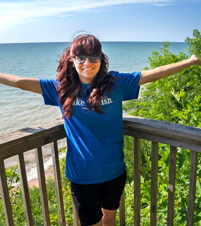 Teenage girl wearing Make-A-Wish shirt holds hands up in the air on deck overlooking Lake Michigan.
