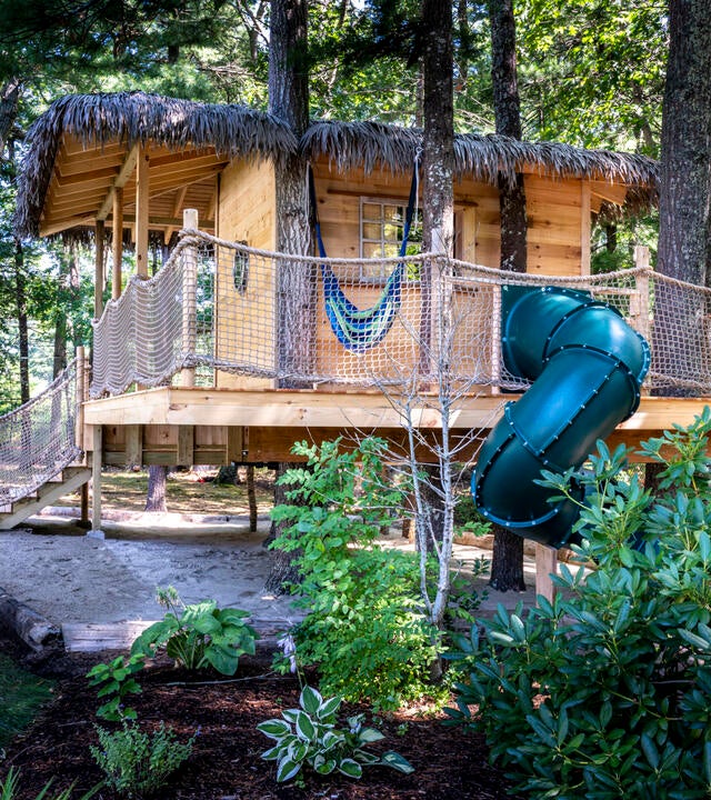Libby's wooden treehouse nestled among the trees, with stairs and a thatched roof and a green twisty slide