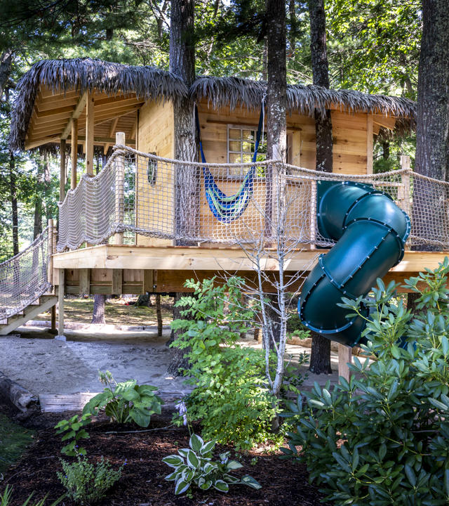 Libby's wooden treehouse nestled among the trees, with stairs and a thatched roof and a green twisty slide