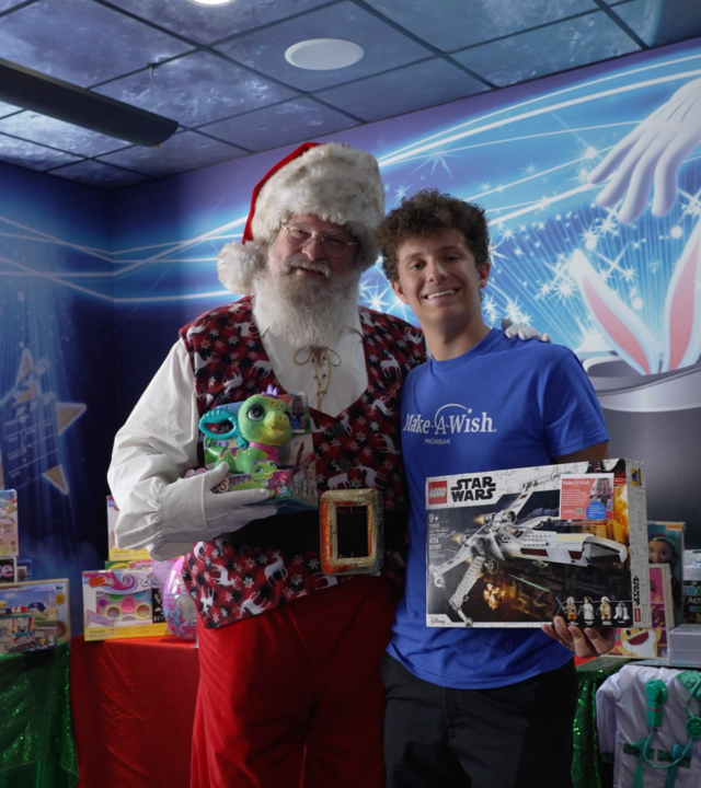 Wish kid in blue shirt standing with Santa in front of toys for Christmas