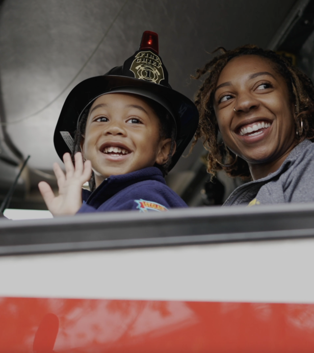 Wish kid in fireman coat and hat with mom waving inside firetruck
