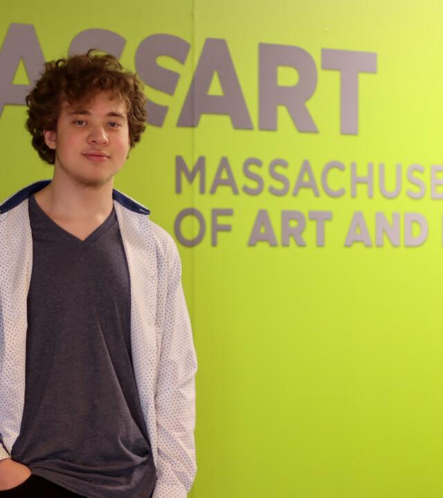 Wish recipient Nate standing in front of the MassArt logo on a bright green wall