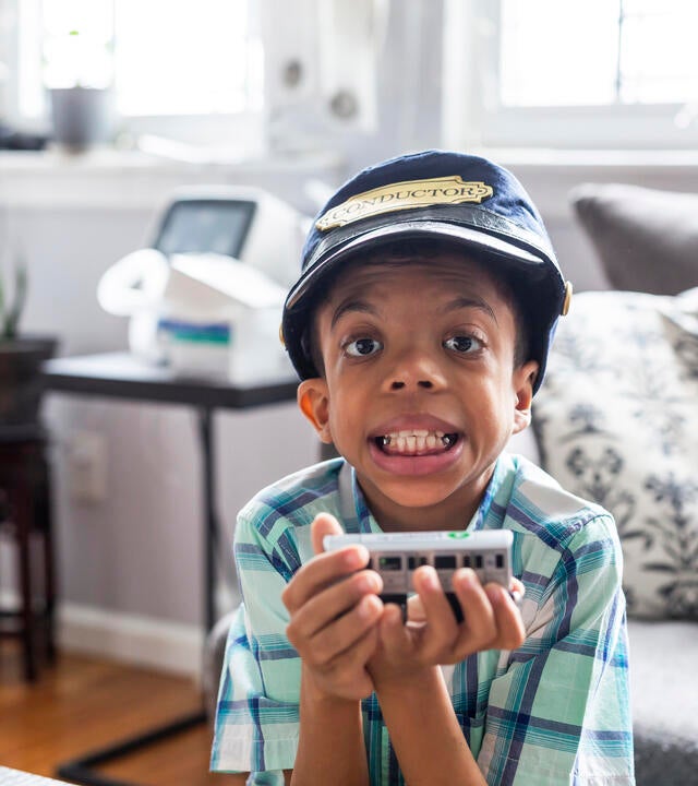 Wish kid Hazael sitting in living room holding toy train with conductor's hat on head