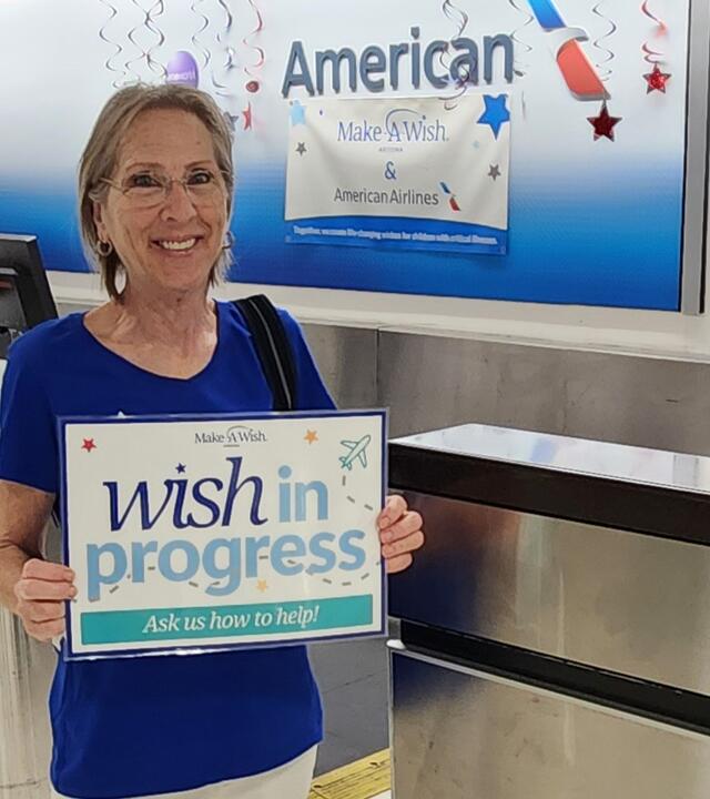 Janet DeYoung standing in the airport with "Wish In Progress" sign