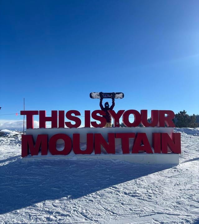 girl on snowy mountaintop holding snowboard above her head while standing behind a sign that reads, THIS IS YOUR MOUNTAIN
