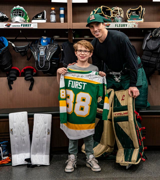 Cameron holding up a custom jersey next to Marc Fleury in the Wild locker room