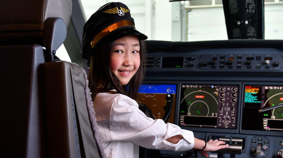 A child with long straight black hair sits in the pilot's seat of an airplane, surrounded by panels of video screens, buttons and levers. The child is looking back over a shoulder at the camera and smiling. The child is wearing a white long sleeved shirt and a black pilot's hat adorned with a red band and golden wings.