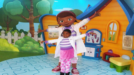 An 8-year-old child in a purple, pink, and white dress, white lab coat, and pink and purple polka dot leggings poses with Doc McStuffins in front of an orange and blue cartoon playhouse stage set.