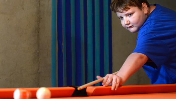 A child with short brown hair wearing a royal blue Make-A-Wish t-shirt lines up a shot on a striped orange billiards ball. Behind the child is a gray concrete wall hung with streamers in two shades of blue.