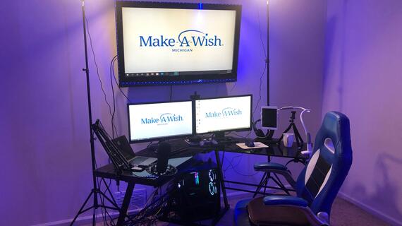 A streaming set-up with three monitors, microphone, and various other technology. The room is lit in soft purple light.