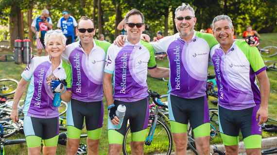Five adults pose with their arms around each other. They are wearing matching green and purple cycling outfits with the Trinity Health logo. In the background, their bikes are laying on the grass.