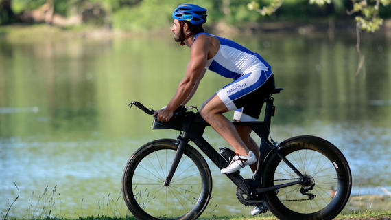 An adult wearing a blue and white cycling outfit and royal blue helmet rides a black bike along a waterside trail.