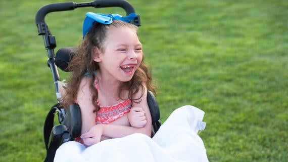 Sitting in a wheelchair with a white lap blanket and a big blue bow tied in curly hair, a 6-year-old child laughs with unrestrained joy. The background is a field of smooth green grass.