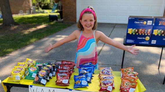 A child in a hot pink headband and brightly colored dress stands with arms thrown out wide behind a snack stand fundraiser. In front of the child, a table covered with a bright yellow plastic tablecloth is filled with individual variety bags of potato chips. A sign taped to the front of the table reads "WAM (Wish-A-Mile) Fundraiser" and advertises lemonade, water, and snacks for sale.