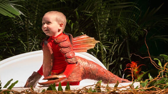 A 4-year-old child wears a red Spinosaurus costume while exploring an area decorated to look like a jungle. Directly behind the child is an oversized model of a dinosaur egg.