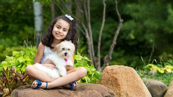 A 7-year-old child with shoulder-length curly dark hair cuddles a fluffy white puppy while sitting cross-legged on a large rock. The child is wearing a white lace tank top and shorts with blue sandals. In the background there are large green plants and trees.