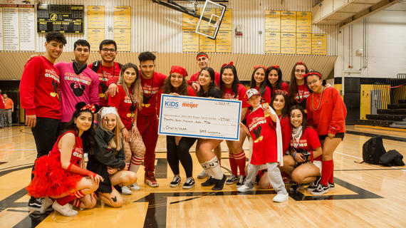 17 high school students wear red headbands and shirts that read "Sterling Heights Clubhouse" as they pose in their school gymnasium with a child. The group holds an oversized check for $27,060 dollars made out to Make-A-Wish Michigan.