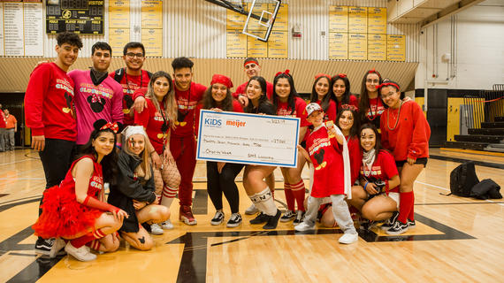17 high school students wear red headbands and shirts that read "Sterling Heights Clubhouse" as they pose in their school gymnasium with a child. The group holds an oversized check for $27,060 dollars