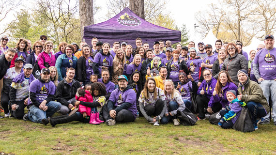 A group of about 60 people of varying ages pose for a group photo. Nearly all of them are wearing purple t-shirts with the Planet Fitness logo and text that reads "The world judges. We don't." A purple pop-up tent with the Planet Fitness logo is behind the group of people.