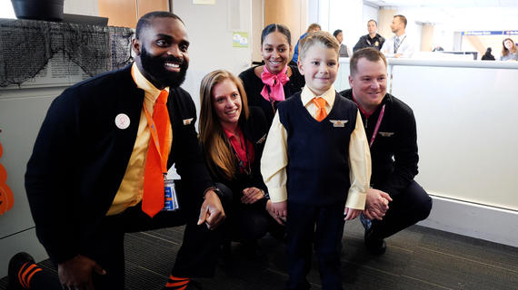 A six-year-old child with short blond hair poses for a photo with the flight crew for a wish trip. The child wears a custom matching flight attendant's uniform, complete with a pair of wings pinned to a navy blue vest. Four adults in navy suits with pale yellow shirts and orange ties or scarves kneel behind the child in the airport boarding area. 