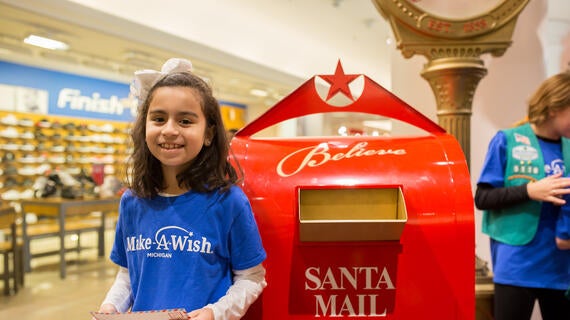 A child with dark curly hair held in place by a big white bow stands beside a red mailbox with a star on top that reads "Santa Mail". The child is smiling brightly and wearing a royal blue Make-A-Wish t-shirt over a long-sleeved white shirt. In the child's hands is an enveloped addressed to Santa Claus, The North Pole.