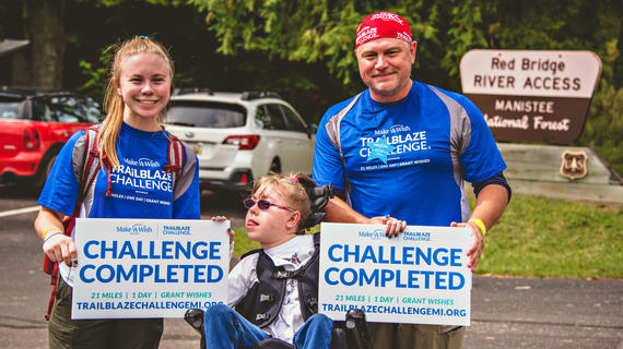 A teenager with pink glasses and a messy blond ponytail sits in a wheelchair between their twin and father, who are both wearing royal blue shirts with the Trailblaze Challenge logo and holding signs that read "Challenge Completed".