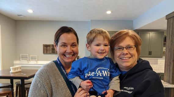 A young child with short blond hair is wearing a royal blue Make-A-wish t-shirt over a long-sleeved gray shirt and clutching a stuffed Mickey Mouse toy as two wish-granting volunteers hug him during a wish discovery meeting.
