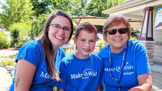 A child with short light brown hair sits at an outdoor table between two wish granting volunteers during a wish reveal celebration. All three are wearing royal blue Make-A-Wish t-shirts. Behind them is a well landscaped garden with colorful flowers and de