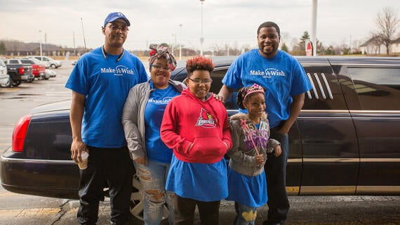 A family of five pose in front of a black limo parked in a mall parking lot. The family is wearing royal blue Make-A-Wish t-shirts, jeanA family of five pose in front of a black limo parked in a mall parking lot. The family is wearing royal blue Make-A-Wish t-shirts, jeans, and hoodies or zipped jackets. The youngest child is also wearing a pis, and hoodies or zipped jackets. The youngest child is also wearing a pi