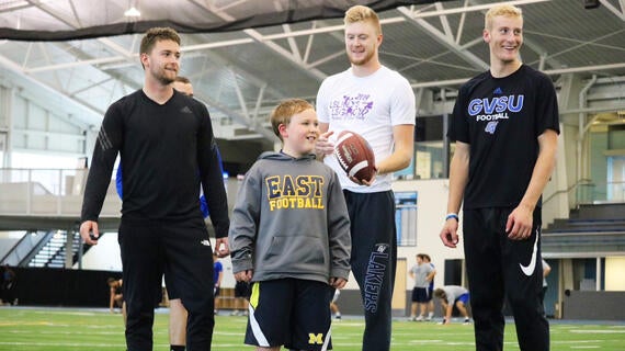 Three college football players pose for a photo on the football field with a young blond child wearing a gray hoodie and black shorts. The player in the middle is holding a football.