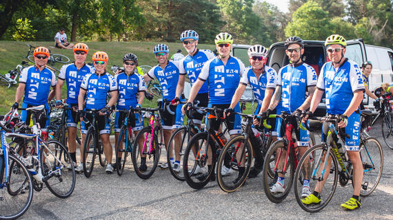 Ten members of the Eaton Wish-A-Mile cycling team pose in a line for a photo with their bikes. They are wearing matching blue and white cycling outfits with the Eaton logo on them and brightly colored helmets. 