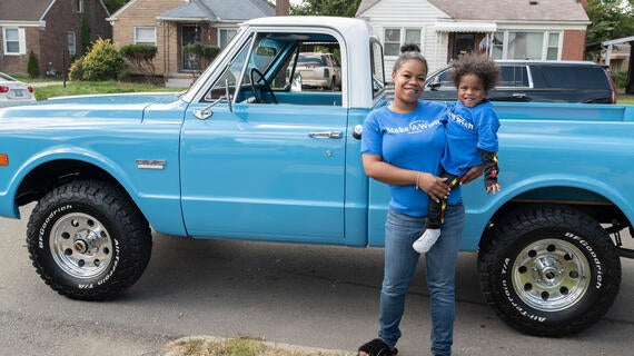 An adult with dark hair pulled into a bun holds a child with dark curly hair in front of a light blue pickup truck. Both are wearing royal blue Make-A-Wish t-shirts.