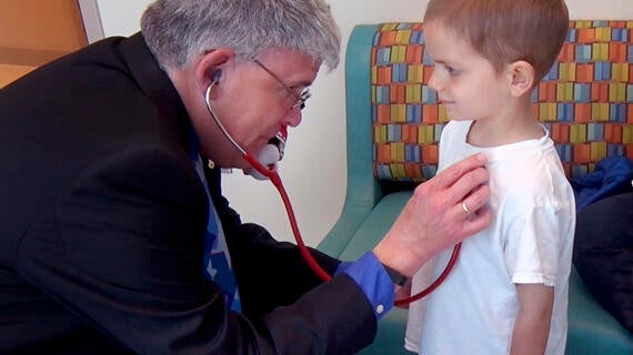 An adult with short gray hair wearing a dark jacket hold a stethoscope to the chest of a young child wearing a white t-shirt.
