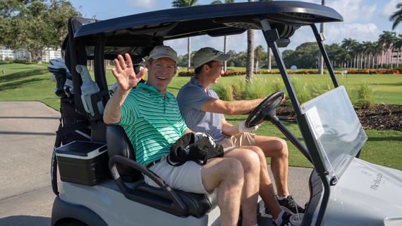 Men on a golf cart waving and smiling on the course.
