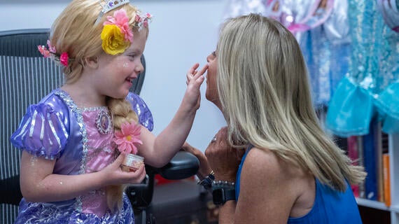 A young girl dressed as a princess putting sparkly glitter on an adult woman's cheek