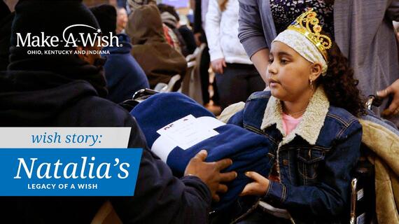 A young girl wearing a crown and denim jacket is handing a blue blanket to someone, in a Make-A-Wish event setting. The girl is in a wheelchair, with the text "Natalia's Legacy of a Wish" overlayed on the image.